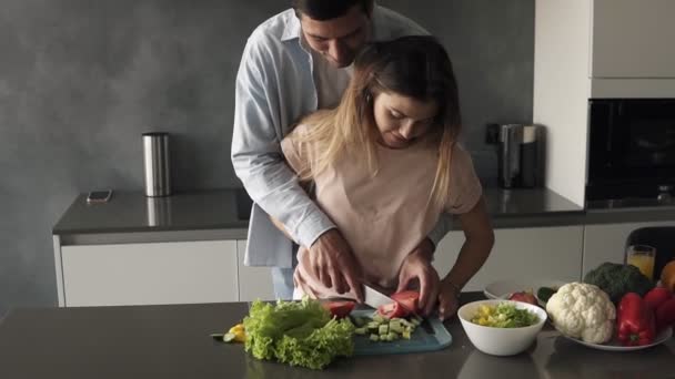 Kærlig par på det grå køkken madlavning sammen. Ung kvinde makulere grøntsager sammen med sin mand. Høj mand står bag og lærer sin kone at skære tomat. Forbered frokost i romantisk – Stock-video