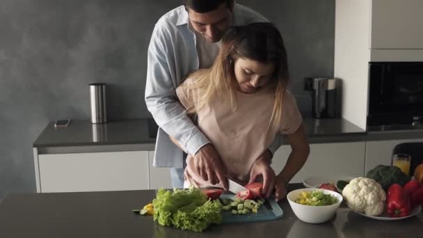 Casal amoroso na cozinha cinzenta cozinhar juntos. Jovem mulher cortando legumes junto com seu marido. Um homem alto atrás de si e ensina a mulher a cortar tomate. Prepare o almoço em romântico — Vídeo de Stock