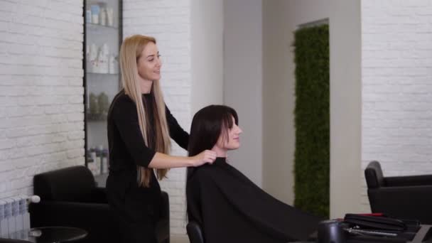 女性の美容師は、長いストレートブラウンの髪で彼女の女性のクライアントに取り組んで終了しました。女性は笑顔と彼女の新しい髪のスタイルについて喜んでいます.美容師保護ケープを削除します。サイドビュー — ストック動画