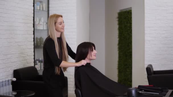 Parrucchiere femminile ha appena finito di lavorare sul suo cliente femminile con lunghi capelli castani lisci. Donna è sorridente e felice per il suo nuovo stile di capelli. Parrucchiere rimozione mantello protettivo. Vista laterale — Video Stock