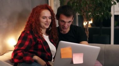 Gri dizüstü bilgisayarlı güzel bir çift kanepede oturuyor. Çift - kısa boylu bir adam ve kızıl saçlı kız birlikte dizüstü bilgisayardan komik bir şey izliyorlar. Konuşurken ve gülümserken. Kanepede oturuyorum.