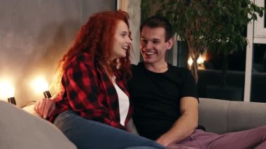 Sevgi dolu, pozitif bir adam ve uzun saçlı mutlu bir kadının evlerindeki kanepede dinlenirken sarılıp sohbet edişinin, arka planda ev bitkisi olan şık oturma odasının görüntülerini kapatın.