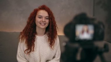 Kızıl saçlı genç kız vlogger 'ın portresi, takipçileri için çevrimiçi blog için kamera önünde gülümseyen ve işaret eden videolar kaydediyor. Kadın kot pantolon ve beyaz kazak giyiyor. Hızlandırılmış