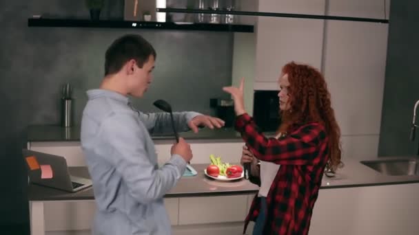 Kaukaski młody mąż rodziny i ruda żona śpiewają w zupie chochla i szczypce makaronu mikrofon razem przygotować za pomocą kuchni jako scena, taniec zabawy słuchać muzyki na kuchni. Powoli. — Wideo stockowe