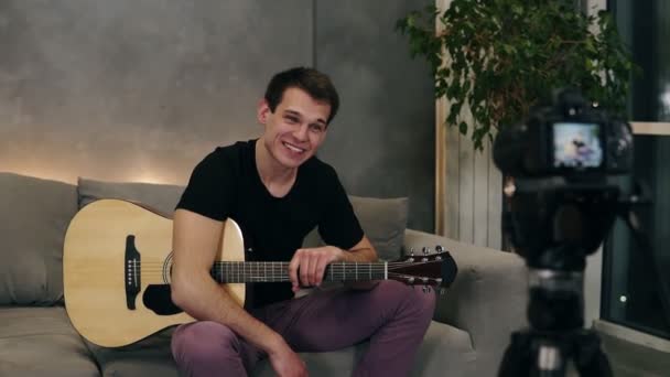 Snygg kaukasisk man i svart T-shirt gör video blogg om musikinstrument eller spela in låten för sina anhängare när du sitter på en soffa hemma. Inspelningskamera på en förgrund — Stockvideo