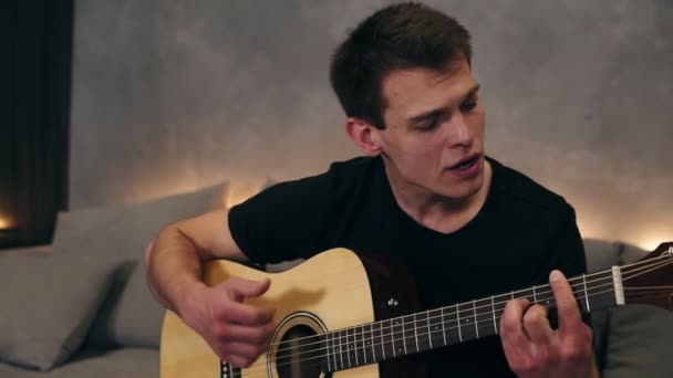 Retrato de um homem alegre cantando uma canção lírica e emocional. Tocar em uma guitarra acústica e apreciá-lo, enquanto sentado em um sofá cinza com uma luz de fundo sob. Movimento lento — Vídeo de Stock