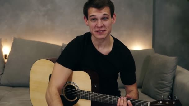 Retrato de hombre caucásico de pelo corto con camiseta negra haciendo video blog sobre instrumentos musicales o grabando la canción para sus seguidores mientras está sentado en un sofá en casa. Mirando a la cámara — Vídeos de Stock