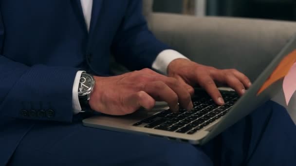 Primo piano delle mani degli uomini d'affari che digitano sulla tastiera del computer portatile mentre sono seduti sul divano con un computer portatile grigio sulle ginocchia. Un uomo vestito blu formale e costoso, orologi di fantasia — Video Stock