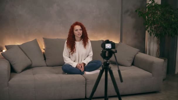 Retrato de una hermosa mujer de pelo largo sentada en un gran sofá gris con retroiluminación en el fondo en la sala de estar interior del loft y hablando con la cámara. Joven blogger haciendo un video — Vídeo de stock