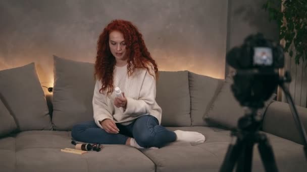 Krullend rood hoofd jong meisje vlogger praat in de voorkant van de camera opname video voor online blog op internet, demosrating cosmetica. De vrouw draagt jeans en een witte trui. Zittend in een woonkamer — Stockvideo