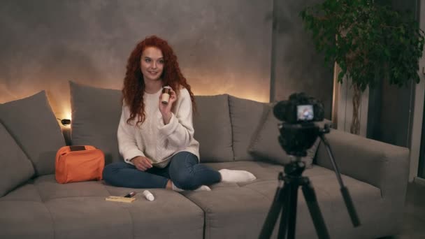 Кучерявий червоний керівник молодої дівчини краси відеоблогер розмовляє перед фотоапаратом для онлайн-блогів в Інтернеті, демонструючи косметику, яка у неї в сумці. Жінка в джинсах і білому — стокове відео