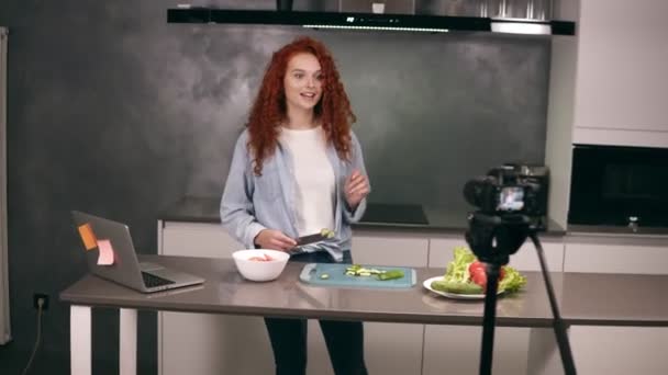 Молодая женщина в повседневной одежде ведет кулинарный блог и записывает видео в сером кухонном баре. Видеосъемка блога - красноголовая резка помидоров на приборной панели и объяснение рецепта. Современные — стоковое видео