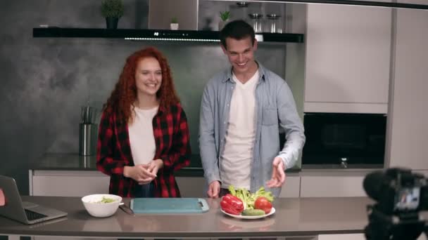 Νεαρό ζευγάρι σε casual μαγείρεμα μαζί και καταγραφή βίντεο τροφίμων blog στην κάμερα στο πατάρι, γκρι κουζίνα στο σπίτι. Ενθουσιασμένοι καλωσορίζουν τους οπαδούς τους, κοιτάζοντας την κάμερα γυρίσματα σε πρώτο πλάνο — Αρχείο Βίντεο