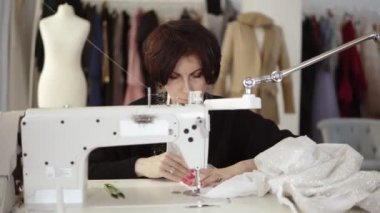 Yetişkin bir kadın terzinin portresi dikiş makinesinde çalışıyor, beyaz bir el yapımı elbise üzerinde çalışıyor. Moda kıyafetleri ve arka planda beyaz manken.