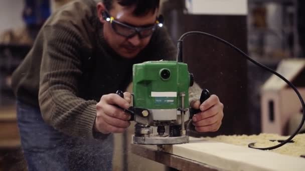 Seorang ahli dalam kacamata pelindung bekerja dengan mesin penggiling manual di bengkel kayu. Menggiling papan kayu besar. Debu dan keripik berserakan di lantai. Gerakan lambat — Stok Video