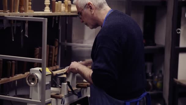 A senior man in eyeglasses standing in front equipment and cutting wooden knob out of wood piece spinning on machine using chisel, close up shot. Slow motion. Side view — Stock Video