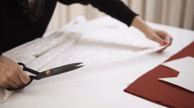 Moda tasarımcısı, terzilik aletleriyle dolu bir stüdyoda beyaz, parlak kumaşla çalışıyor. Desen, makas. Terzilik işyeri. Terzi çizim çizgilerindeki gelinlik detaylarını kesiyor.