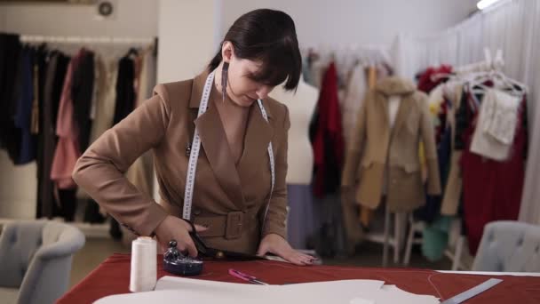 Diseñadora de moda femenina en elegante abrigo marrón y pendientes que trabajan con tela roja en el estudio lleno de herramientas de sastrería - patrón, tijeras, cinta métrica. Lugar de trabajo de costurera. Cortes a medida — Vídeos de Stock