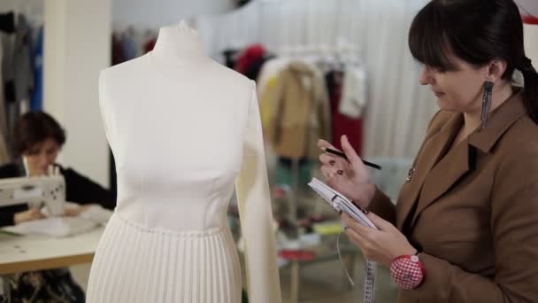 Moderno estudio de costura, morena concentrada diseñadora de moda femenina después de medir a partir de un traje blanco en maniquí tomando notas sosteniendo su cuaderno. En cámara lenta. Vista lateral — Vídeo de stock