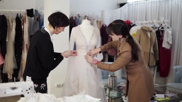 Retrato de dos diseñadoras creando un vestido de novia por orden exclusiva cosiendo telas y fijando detalles en un maniquí. Producción de vestidos de novia. Pequeño negocio — Vídeos de Stock