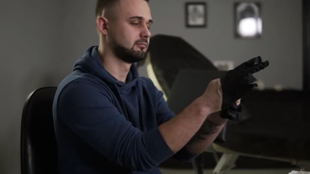 Retrato de un artista del tatuaje poniéndose los guantes negros de látex antes de una sesión de tatuaje en la tienda de tatuajes. Tatuaje sentado en silla en salón gris paredes habitación — Vídeo de stock