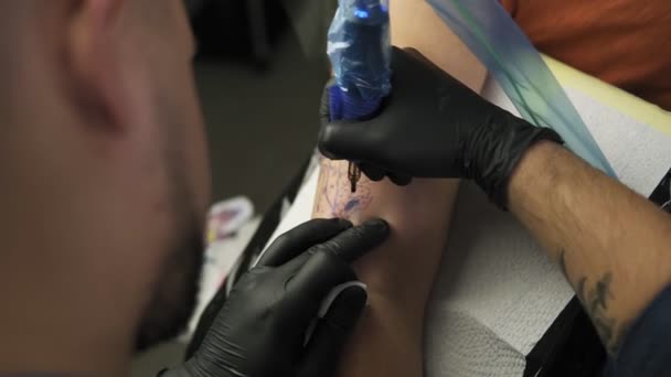 En närbild av en tatueringskonstnär gör en tatuering på en kvinnas arm. En hög upplösning. Professionell tatuerare arbetar i studio. Högvinkelfilm från mästarens axel — Stockvideo