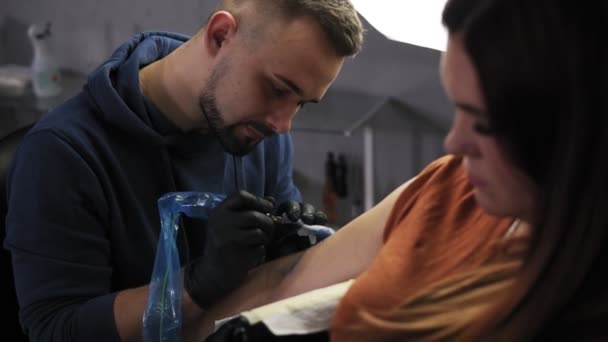 Портрет татуировщика демонстрирует процесс нанесения цветной татуировки. Современный тату-салон. Бородатый татуировщик и клиентка на переднем плане в салоне красоты — стоковое видео