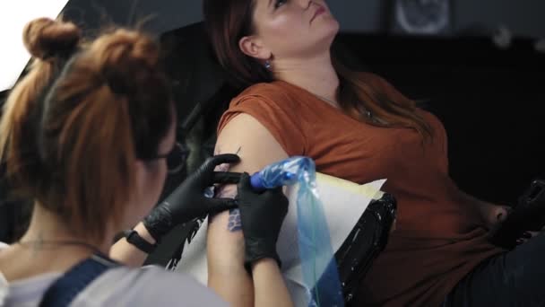 The tattoo artist at work. A red headed girl holding a tattoo machine and applied in a pattern on the skin of her female client. Blue ink in the tattoo machine. High angle footage from the shoulder — Stock Video