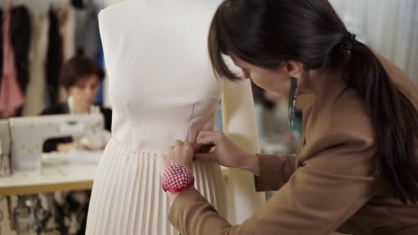 Los diseñadores de moda creativos están trabajando juntos en el estudio de sastrería. Mujer de pelo largo que fija la falda blanca en el maniquí usando alfileres en su almohada de la mano en la muñeca. Mujer trabajando en la máquina de coser — Vídeo de stock