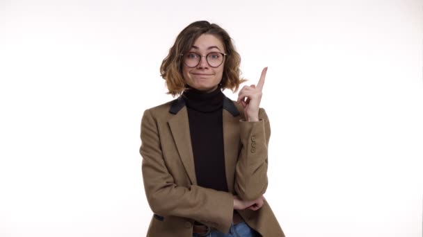 Mujer bonita europea con gafas elegantes y chaqueta marrón se ve confundido y reflexivo, a continuación, se hace una idea. Hermosa joven se emociona después de recordar algo, apuntando hacia arriba con un dedo — Vídeo de stock