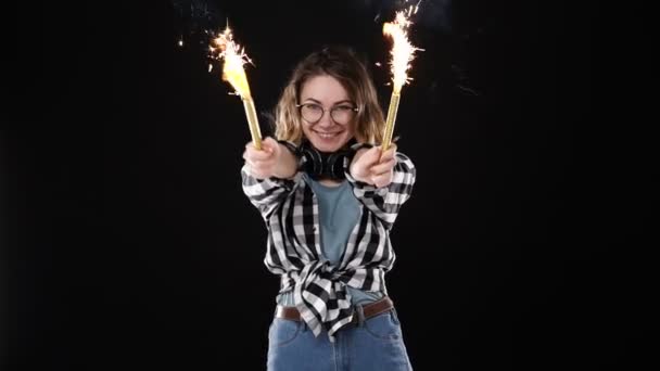 Ευρωπαϊκό hipster κορίτσι σε μαύρο και άσπρο καρό πουκάμισο, ακουστικά στο λαιμό γιορτάζει με sparklers και bengal φωτιά. Ευτυχισμένη νεαρή γυναίκα που απολαμβάνει και διασκεδάζει με μαύρο φόντο. Αργή κίνηση — Αρχείο Βίντεο