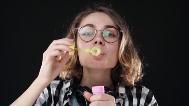 Primer plano retrato de la chica emocional feliz soplando un montón de pompas de jabón con palo sobre fondo negro. Imágenes de estudio de morena con pelo corto y labios sensuales con auriculares en el cuello. Lento. — Vídeo de stock