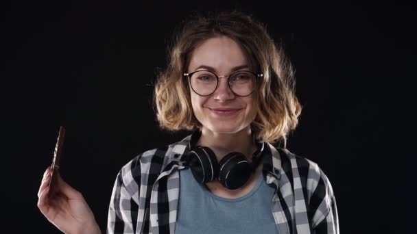 Fermer jeune femme brune avec des lunettes et des écouteurs posant de façon ludique manger tenir dans la main barre de chocolat avec des noix isolées sur fond noir en studio. Personnes émotions sincères concept de style de vie — Video