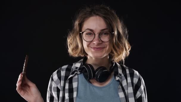 Närbild brunett ung kvinna med glasögon och hörlurar poserar lekfullt äta grepp i handen choklad bar med nötter isolerade över svart bakgrund i studio. Människor uppriktiga känslor livsstil koncept — Stockvideo