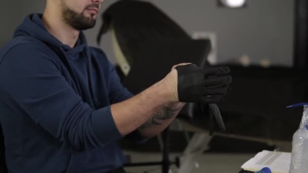 Close up of hands tattoo artist putting on the latex black gloves before a tattoo session at the tattoo shop. Tatuagem sentada na cadeira na sala de paredes cinza salão. Fundo desfocado. Movimento lento — Vídeo de Stock