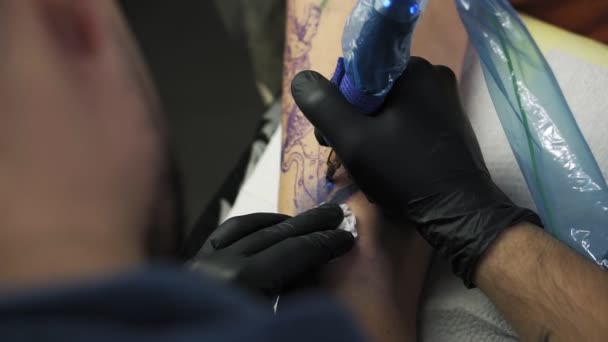 A close-up of a tattoo artist holding tattooing gun and makes a tattoo on a womans arm using blue paint. Professional tattooist works in studio. High angle footage from the shoulder of master — ストック動画