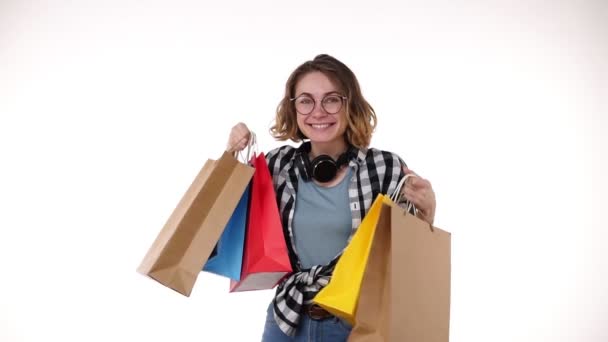 Fröhlich schöne europäische junge Frau mit Brille und Kopfhörer am Hals, bunte Einkaufstüten im Saisonverkauf haltend, fröhlich auf ausgestreckten Händen auf weißem Hintergrund — Stockvideo