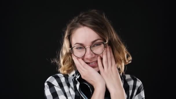 Nahaufnahme Porträt eines jungen europäischen Mädchens, das demütig vor schwarzem Hintergrund in die Kamera lächelt. fröhlich lächelnd, peinlich berührt. Brille und Kopfhörer am Hals tragen — Stockvideo