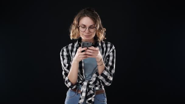 평범 한 옷차림 - 격자무늬 셔츠와 청바지를 입고 스마트폰을 들고 문자 메시지를 입력하거나 채팅을 하는 유럽 여성의 사진 - 이 검은 배경에 고립되어 있었다. 감정의 개념 — 비디오