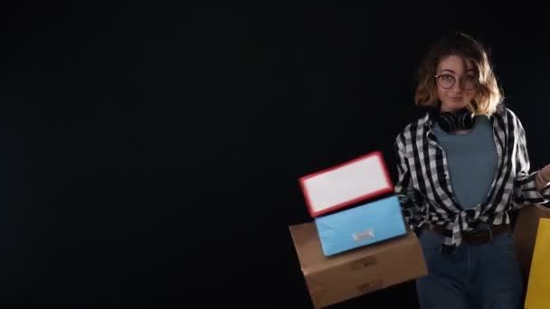 Hermosa mujer sonriente en camisa a cuadros y jeans caminando felizmente con coloridas bolsas de compras, posando juguetonamente sobre fondo negro. Usando gafas y auriculares negros en el cuello. Movimiento lento — Vídeo de stock