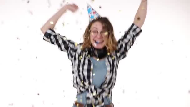 Cheerful, excited young woman in plaid shirt and headphones on neck in birthday colorful hat blow in pipe and dancing, falling confetti isolated over white background in studio. People sincere — Stok video