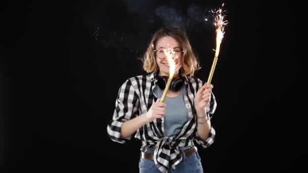 Ragazza hipster europea in camicia scozzese in bianco e nero, cuffie sul collo che festeggiano con scintille e fuoco del bengala. Giovane donna felice divertirsi e giocosamente ballare sullo sfondo nero. Lento. — Video Stock