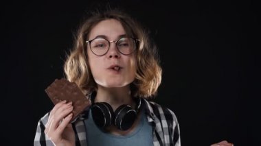 Close up brunette young woman with glasses and headphones posing playfully eat, chewing chocolate bar with nuts isolated over black background in studio. People sincere emotions lifestyle concept