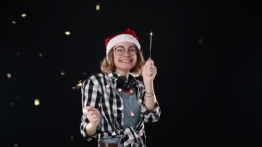 Noel Baba şapkalı, renkli çelenkli ve boynunda iki maytap olan sevimli bir kadın Noel partisinde siyah arka planda dans ediyor. Gülümseyen, neşeli genç kadın eğleniyor.