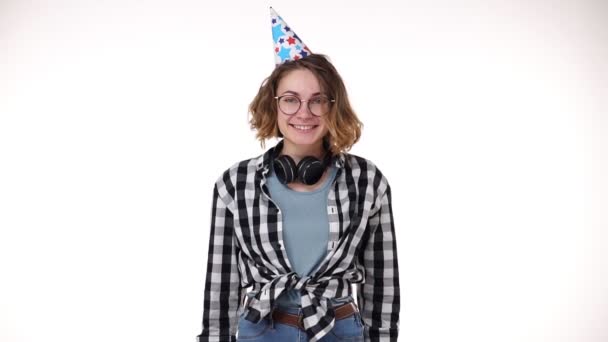 Portrait of excited young woman in plaid shirt and headphones on neck in birthday colorful hat blow in pipe, falling confetti isolated over white background in studio. People sincere emotions, holiday — 비디오