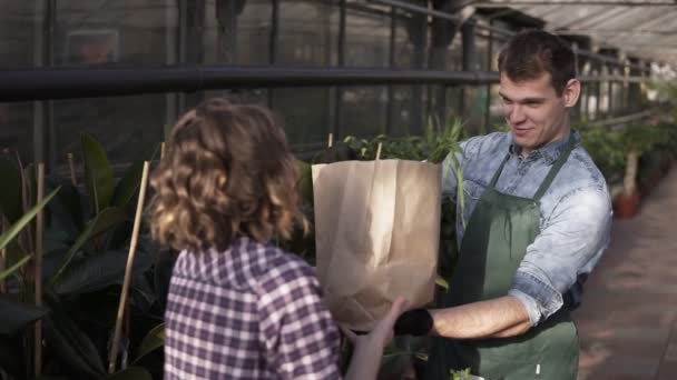 Positive,tall salesman in green apron giving fresh vegetables in paper bag to customer in greenhouse market. Smiling man selling vegetables to female customer. People and healthy lifestyle concept — 비디오