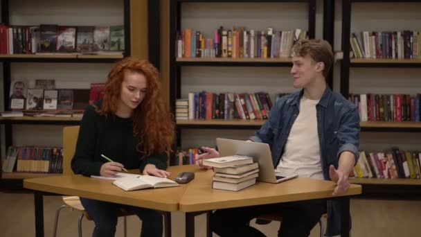 Молодые студенты мужского и женского пола разговаривают с помощью ноутбука, объясняя работу курса в библиотеке. Активно обсуждаем что-то сидя за деревянным столом. Вид спереди — стоковое видео