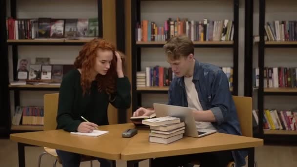 若い男性と女性の学生が話し、図書館でのコースワークの勉強を説明します。木のテーブルに座って本と積極的に何かを議論する。正面図 — ストック動画