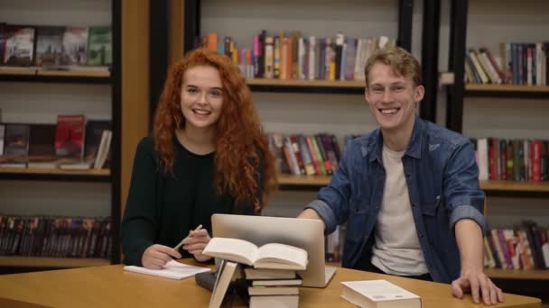 Dos jóvenes estudiantes europeos, hombres y mujeres, sentados a la mesa con libros y portátil en la biblioteca preparándose para los exámenes. Estanterías en la biblioteca con muchos libros en segundo plano — Vídeos de Stock