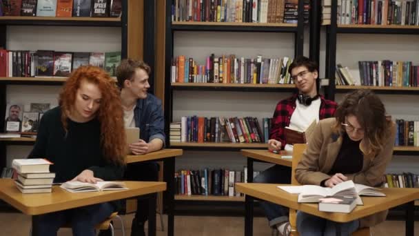 Intelligente vrouwelijke en mannelijke studenten leren, de voorbereiding van examens in de universiteitsbibliotheek zitten aan de afzonderlijke tafels over boekenplank achtergrond. Charmante jongeren leren in de bibliotheek — Stockvideo